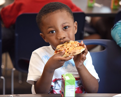A boy taking eating a piece of pizza and has a juice box sitting on the table in front of him.