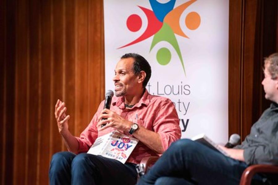 Award-winning poet and essayist Ross Gay sitting in a chair, holding a microphone while discussing his book "Inciting Joy”