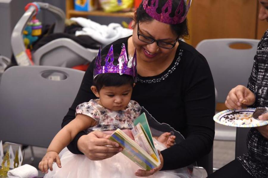A smiling woman with glasses reads a board book to her little girl who is wearing a pink flowered dress and a purple crown and sitting on her lap