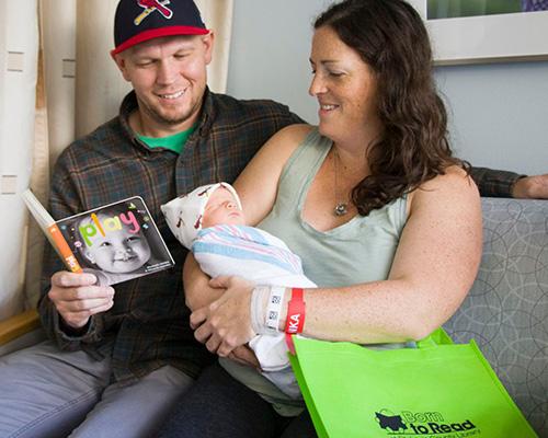 A woman holds her newborn baby while her husband who has his arm around her reads a board book included in a gifted Born to Read bag